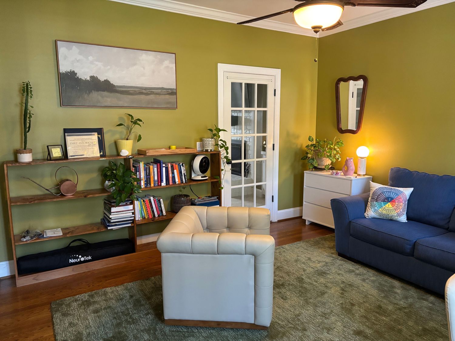 The Third Space Psychotherapy office in Whitsett, NC: a warm room with olive-green walls, a blue sofa, bookshelves, plants, and soft light